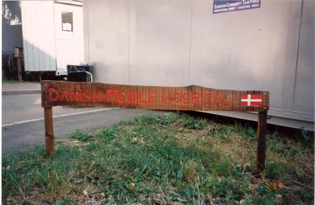 Hand-painted wooden sign reading Danish Mobile Hospital with a Danish flag in front of the hospital containers