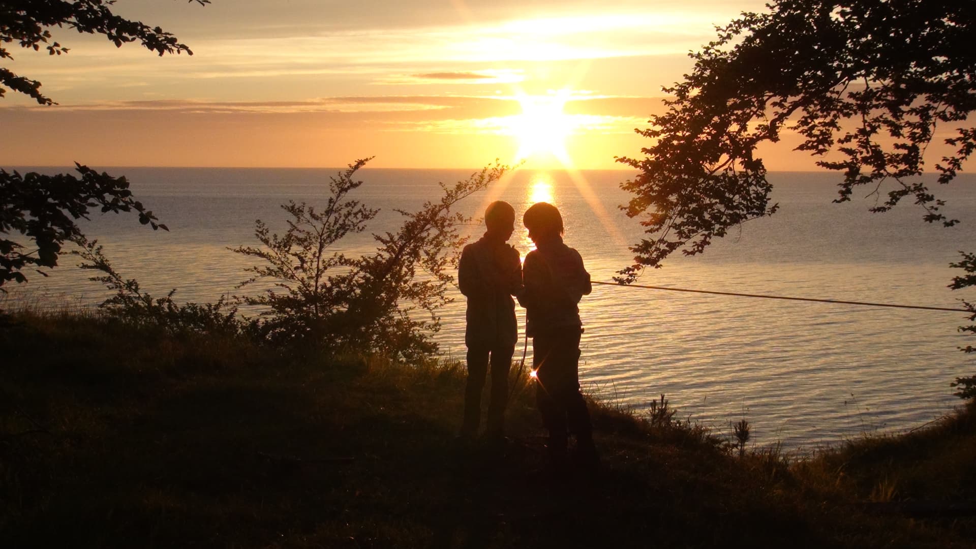 To unge i silhuet på en skrænt ved havet i solnedgang, med et reb udspændt ved siden af