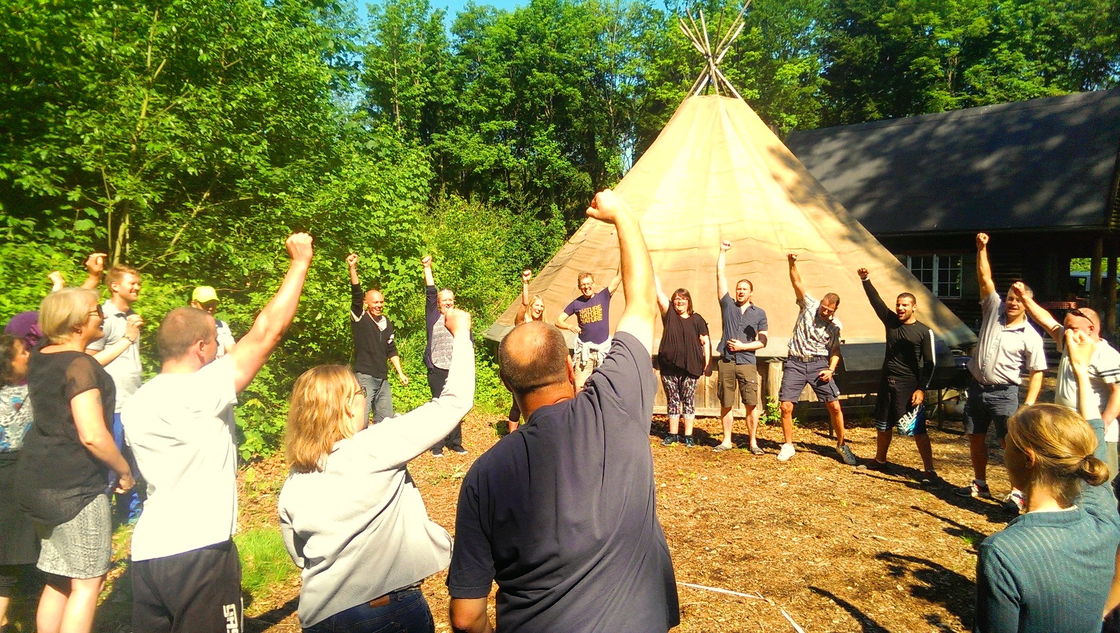 En stor gruppe voksne står i en rundkreds foran en tipi og løfter armene sammen